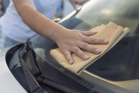 A girl in a white t-shirt wipes the window in the car with a yellow special window cloth.Close-up of a hand and a rag.Work and cleanliness concept.の写真素材
