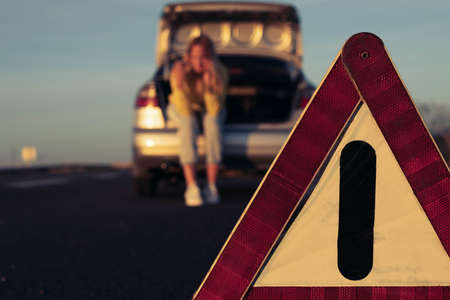 a girl in casual clothes sits in the trunk, in the foreground is an emergency stop sign close-up and in focus, the girl is blurred in the background. Car breakdown on the highway, on the roadの写真素材