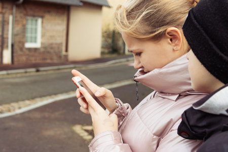 a teenage girl of European appearance with blond hair in a pink jacket and a boy of European appearance in a black hat and black jacket, stand on the street and look at the phone,の写真素材