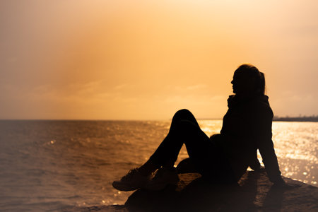 silhouette of a girl sitting on a stone on the sea at sunset, close-up on the silhouette there is a place for an inscriptionの写真素材