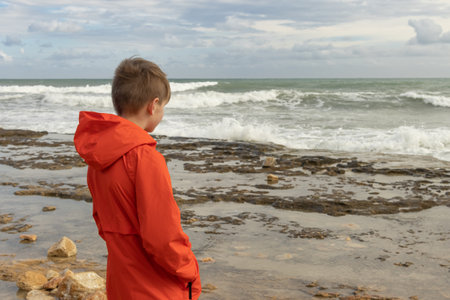 a teenager in an orange windbreaker stands on the beach with his back to the camera and looks at the sea, there is a place for an inscription. Seascapeの写真素材