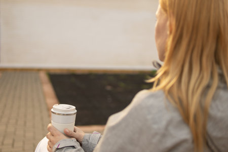 back photo of a girl who is sitting on a bench in the park holding a white thermo mug for tea or coffee in her hands. There is a place for an inscription, a close-up on her hands and a flask.の写真素材