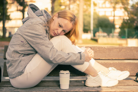 a girl with blond hair sits on a bench with her feet and lowered her head to her feet, the girl sits in a gray jacket and white sweatpants and sneakers, there is a white thermo mug nearbyの写真素材