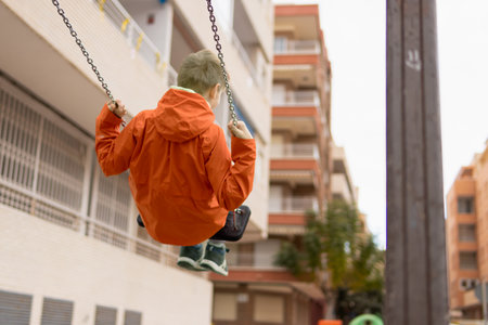 a boy with blond hair in an orange jacket rides on a swing turned with his back to the camera on the street. There is a place for an inscriptionの写真素材