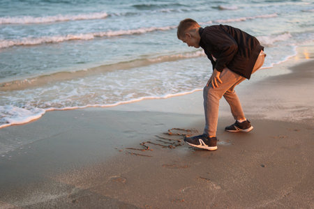a happy child of European appearance sits on the beach near the sea draws on the sand. A child on vacation sits near the seaの写真素材