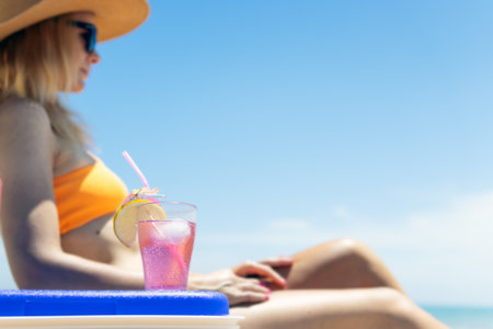Happy young european girl drinks a cocktail and rests in a sun lounger on the beach, space for an inscription portable blue refrigerator by the seaの写真素材