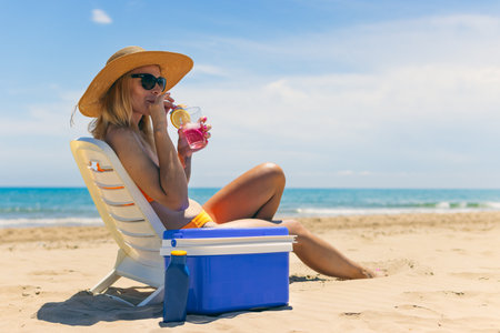 Happy young european girl drinks a cocktail and rests in a sun lounger on the beach, space for an inscription portable blue refrigerator by the seaの写真素材