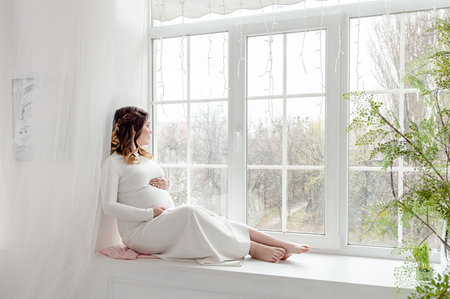 Pregnant girl in a white dress sits on the windowsill and looks out the windowの写真素材