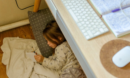 Cute happy little kid girl drawing with pencils lying on warm floor under the table, smiling preschool child daughter enjoy creative activity while parents relaxing on couch at homeの写真素材