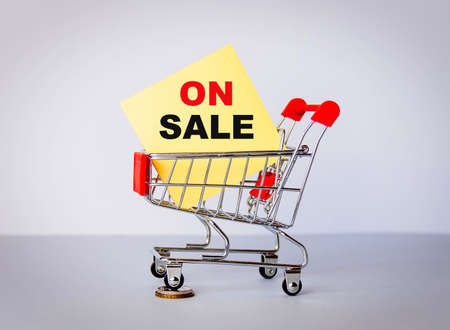 Close-up of a shopping trolley with one wheel on coins with the words ON SALE on a yellow stick.の写真素材
