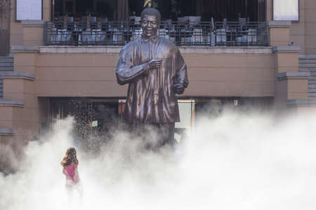 A little girl walking up to the statue of Nelson Mandela while appearing to be emerging out of a thick white fog created by the water sprayed by a nearby fountain. It was a hot summer day in Nelson Mandela square, located in Sandton, South Africaのeditorial素材