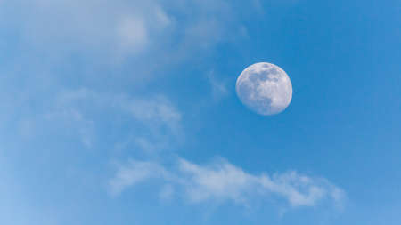 An image of the moon during the day with a clear blue sky and scattered clouds in the backgroundの写真素材