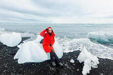 A female tourist in a red jacket and glasses sits on a large piece of ice on Diamond Beach. JÃ¶kulsÃ¡rlÃ³n glacial lagoon. Iceland. Travel through Icelandの写真素材