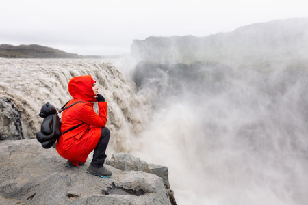 A girl in a red jacket and with a backpack sit and look at the Dettifoss waterfall.の写真素材
