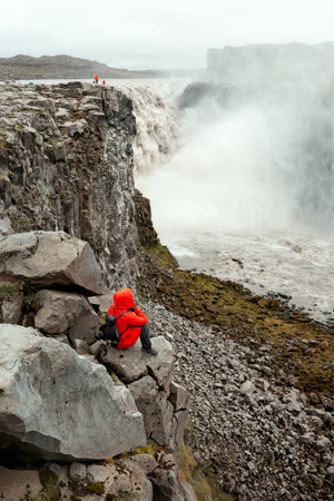 A girl in a red jacket and with a backpack sits on a rock and looks at the Dettifoss waterfall. Icelandの写真素材