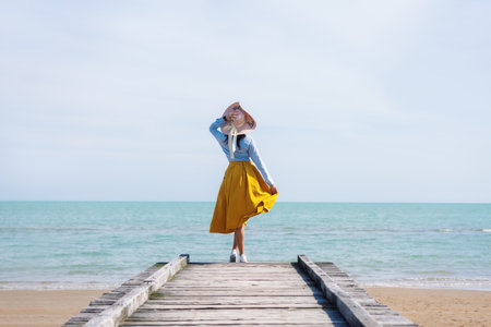 A girl in a hat and a yellow skirt stands on a wooden pier against the background of the sea. Rest at the sea.の写真素材