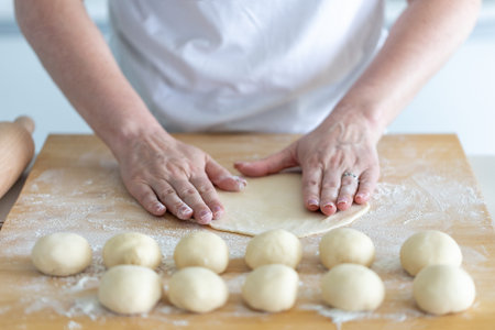 A person rolls out dough on a board, dough blanks are lying nearby.