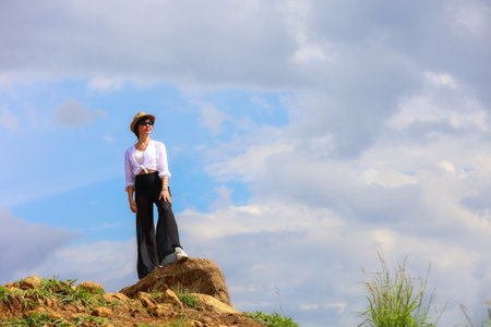 A female tourist in a hat stands on the top of a mountain against a background of a sky with clouds.の写真素材