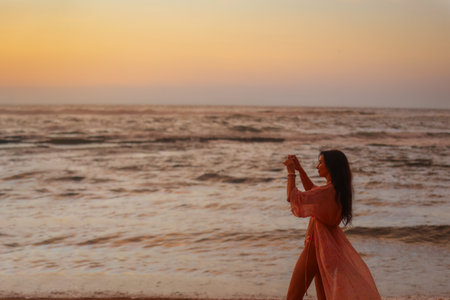Woman taking a photo of sunset on a calm beach with her phoneの写真素材