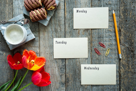 Three colorful tulips on a wooden table. Cup of coffee, cookies. The leaves of the diary Monday...