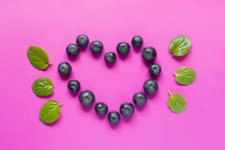 Blueberries laid out in the shape of a heart with mint leaves on bright crimson background.