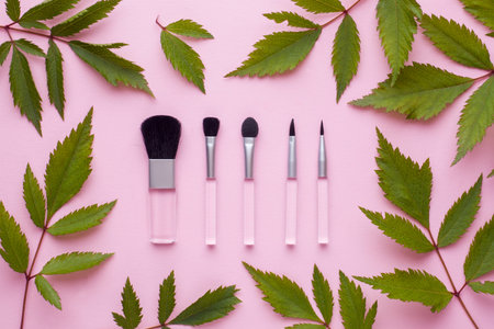 Makeup brushes on a pastel pink background with plant leaves. Beauty concept