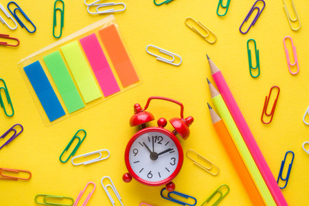 Pencils and colored paper clips, clock alarm on yellow background. Copy space.