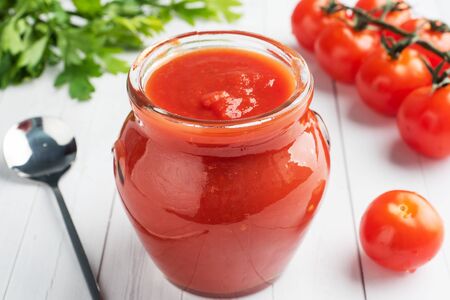 Tomatoes in their own juice or Tomato paste in a glass jar and fresh tomatoes on a white wooden table.の写真素材