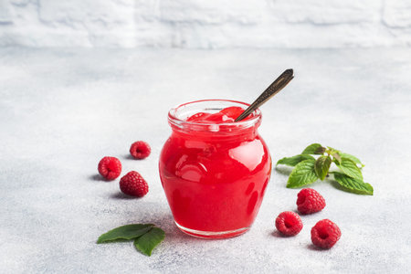 Homemade raspberry jam in a glass jar and fresh raspberries with mint on a gray concrete background....