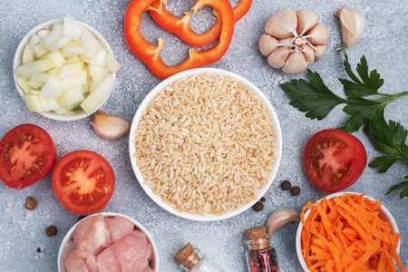 Raw ingredients for cooking pilaf, Brown rice and chicken fillet, grated carrots and chopped onions, pepper tomato garlic herbs and spices. Gray background, top viewの写真素材