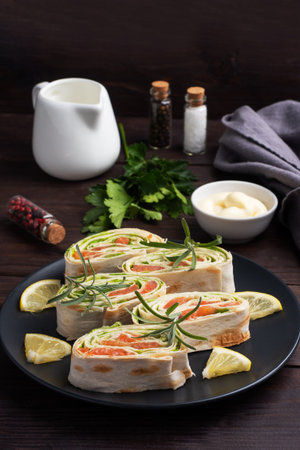 Rolls of thin pita bread and red salted salmon with lettuce leaves on a black ceramic plate, dark...