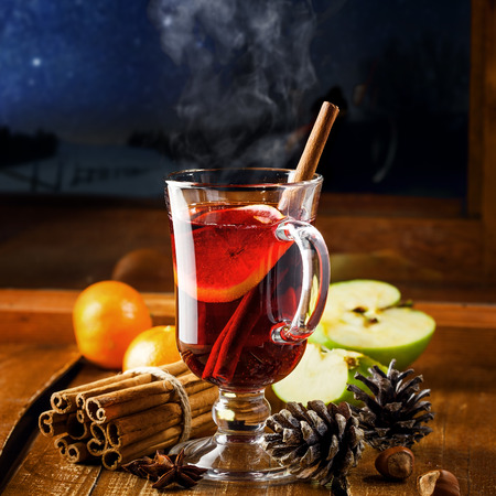 Traditional winter holiday alcoholic drink. Mulled wine with lemon slice on a table. Starry night winter landscape in a window.の写真素材