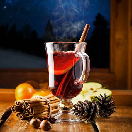 Mulled wine with citrus fruits and cinnamon on a table. Traditional winter hot drink for Christmas Eve party by window. Starry night winter landscape on background.の写真素材