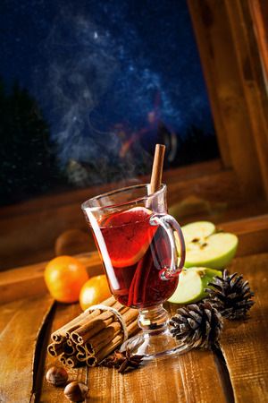 Mulled wine with citrus fruits and cinnamon on a table. Traditional winter hot drink for Christmas Eve party by window. Starry night winter landscape on background.の写真素材