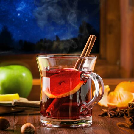 Traditional winter holiday alcoholic drink. Mulled wine with lemon slice on a table. Starry night winter landscape in a window.の写真素材