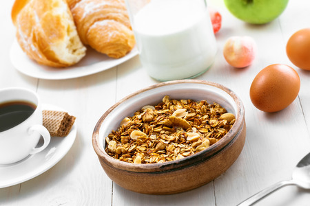 Healthy food for breakfast on a white wooden table. Oatmeal granola with nuts, coffee, croissants, eggs, milk and fruits for delicious healthy meal.の写真素材