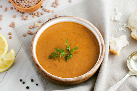 Healthy vegetarian lentil soup in a rustic clay bowl with bread and lemon slices on a white wooden table. Delicious healthy meal served on a canvas tablecloth. Top view shot.の写真素材