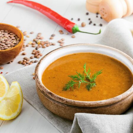 Healthy vegetarian lentil soup in a rustic clay bowl , piece of bread and lemon slices on a white wooden table. Delicious healthy meal served on a canvas tablecloth.の写真素材