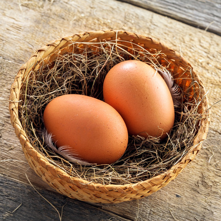 Couple of fresh chicken eggs in a rural basket with hey and feather on a wooden background. Close-up shot. Top view.の写真素材