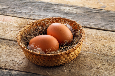 Two fresh farm eggs in a basket with hey and feather on a wooden table. Close-up shot.の写真素材