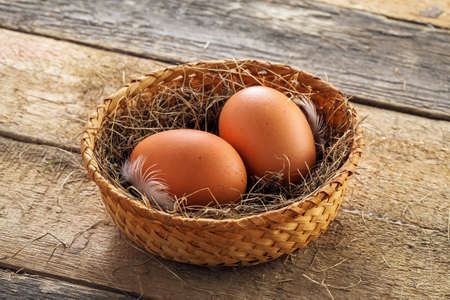 Two fresh farm eggs in a basket with hey and feather on a wooden table. Close-up shot. Top view.の写真素材