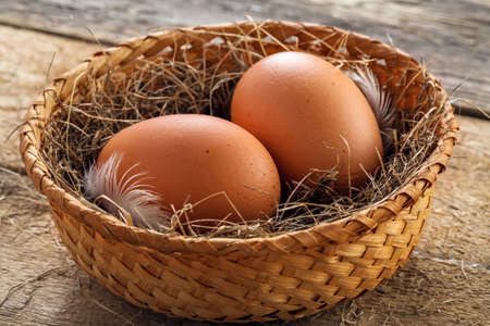 Couple of fresh chicken eggs in a rural basket with hey and feather on a wooden background. Close-up shot.の写真素材