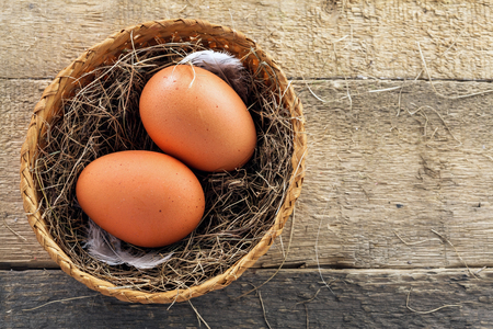 Two fresh farm eggs in a basket with hey and feather on a wooden table. Close-up shot.の写真素材