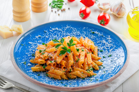 Delicious Italian penne pasta with tomato sauce and parsley on a blue rustic plate. Traditional healthy Italian cuisine meal on a white wooden table.の写真素材