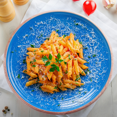 Delicious Italian penne pasta with tomato sauce and parsley on a blue rustic plate. Traditional healthy Italian cuisine meal on a white wooden table. Top view.の写真素材