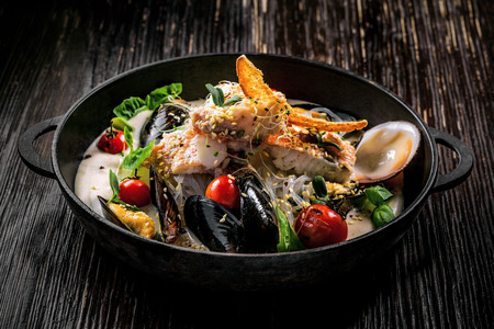 Bowl of delicious seafood meal with rice noodles. Meal made of sea bass fish, mussels, crab meat and other seafood with vegetables on a table.の写真素材
