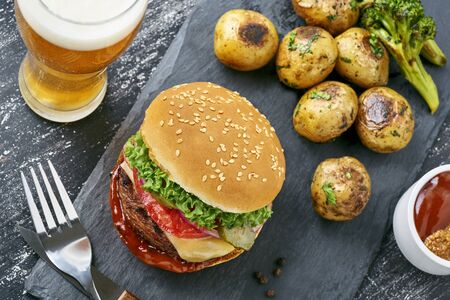 Classic hamburger with tomato, lettuce and onion served with grilled potatoes and broccoli on a black stone board with a glass of beer. Top view, diretly above.の写真素材