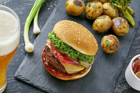 Classic hamburger with tomato, lettuce and onion served with grilled potatoes and broccoli on a black stone board with a glass of beer. Top view, close-up.の写真素材