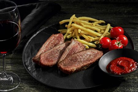 Delicious medium rare steak with French fries, sauce and fresh cherry tomatoes on a black plate. Classic meat meal on a rustic wooden table with red wine glass.の写真素材