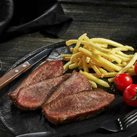 Sliced beef steak with French fries, sauce and cherry tomatoes on a black plate. Delicious American lunch with red wine ona rustic wooden table.の写真素材